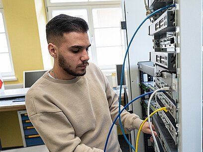 Ein junger Mann steht in einem Serverraum und steckt ein Kabel um.